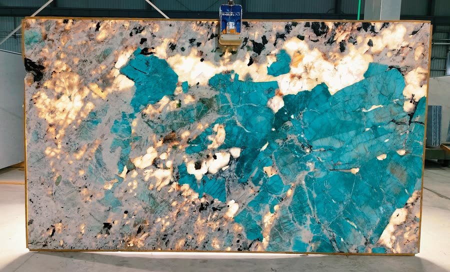 Backlit Amazonite slab standing in a warehouse, lit from behind so turquoise fields and warm white crystalline patches glow through the stone against darker mineral clusters, with a pale border frame around the slab and the warehouse floor visible below.
