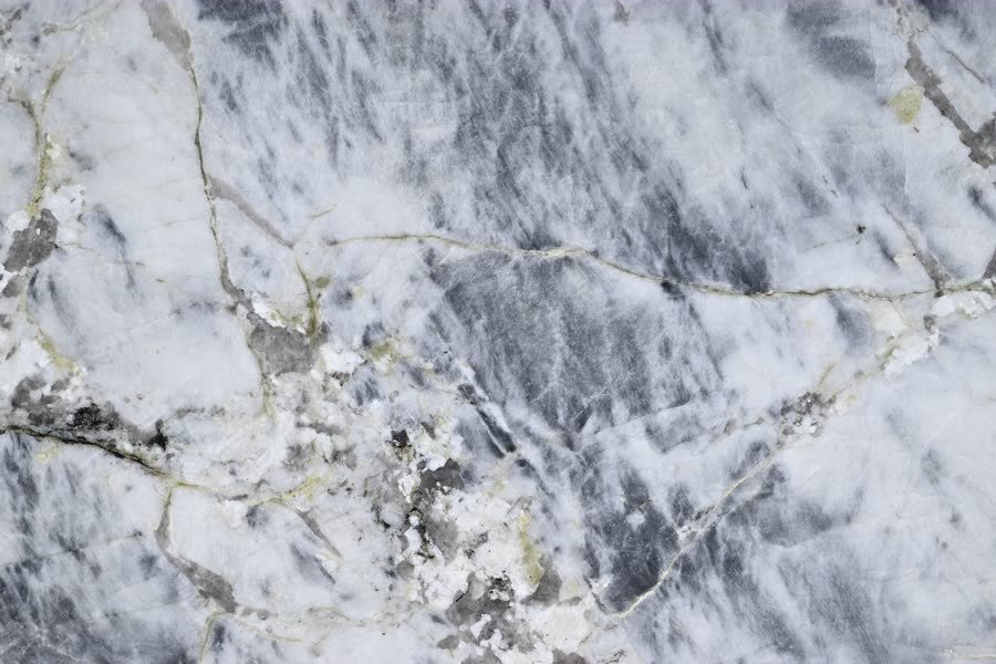 Close-up of a polished Blue Alpine stone surface, a grey-blue and white marbled ground broken by pale crystalline patches and a branching network of thin olive-gold veins.