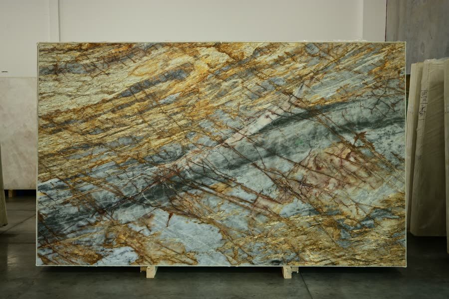 A single large polished stone slab photographed straight on, standing on wooden blocks against a warehouse wall, its surface blue-grey with sweeping gold and amber veining.