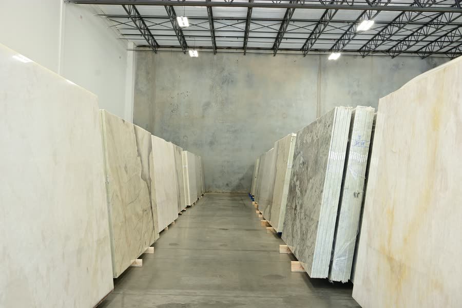 A wide aisle running down the centre of a warehouse between two long rows of upright stone slabs standing on wooden pallets, lit by overhead fixtures under an exposed steel truss ceiling, with a bare concrete wall at the far end.