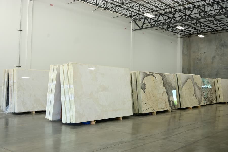 A row of stacked stone slab bundles resting on wooden pallets along the wall of a warehouse, receding to the right, with pale cream slabs in the foreground and darker veined and green-toned slabs further back on a polished concrete floor.