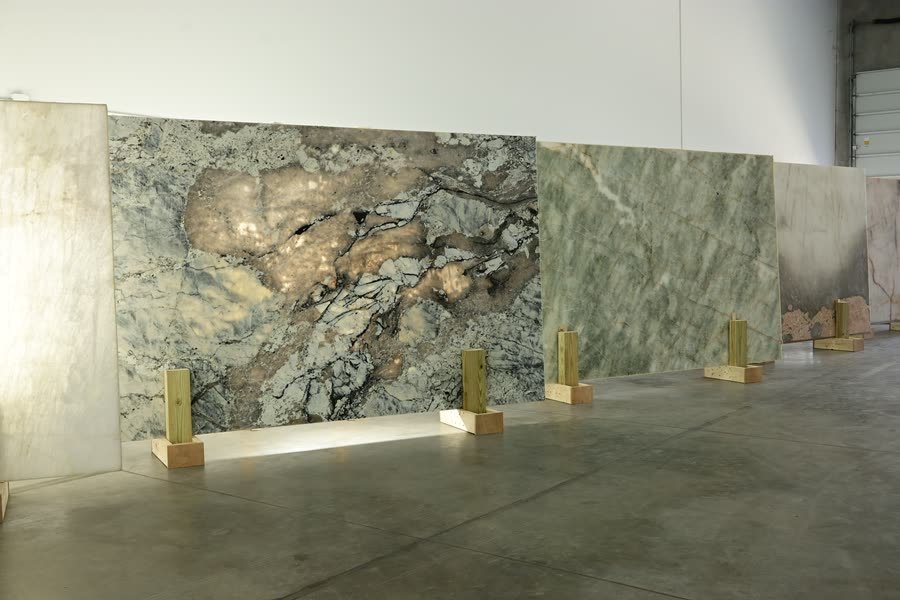 Individual polished stone slabs standing upright on wooden A-frame supports in a row against a warehouse wall: a large grey slab with gold and rust mineral clouding in the foreground, a sage-green veined slab beside it, and a pink-toned slab further along, above a polished concrete floor.
