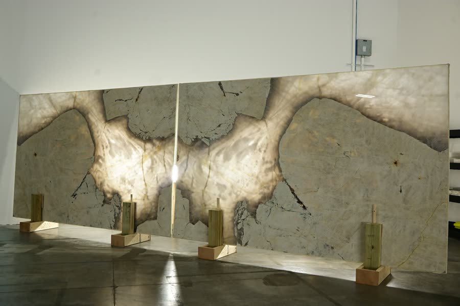 Two large stone slabs standing side by side against a warehouse wall, their pale grey and cream veining mirrored across the central seam to form a symmetrical pattern, supported by wooden blocks on a dark polished concrete floor.
